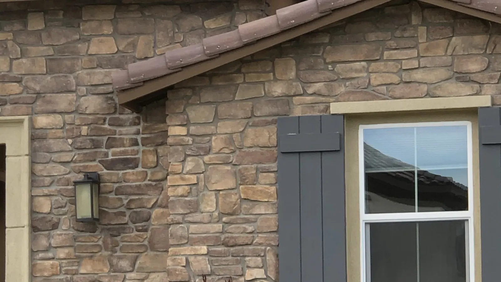 Una fachada de una casa de un solo piso cubierta con chapa de piedra manufacturada, alrededor de una ventana con contraventanas, bajo un techo de teja de barro.